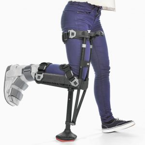 iWalk 2.0 shown on the right leg of a female model wearing jeans. The iWalk is a peg-leg style device; all black with a wide tip. The leg using the iWalk bends at a 90-degree angle and sits in a cradle, while the iWalk base acts as a 'foot.'