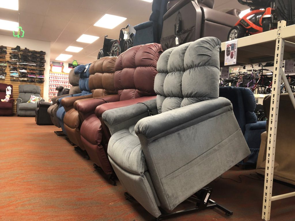 A row of 6 power lift recliners in the up/lift position on Oswald's medical showroom floor. There is a big shelf over the chairs with more chairs, scooters and wheelchairs on display. Picture taken for the best power recliner blog.