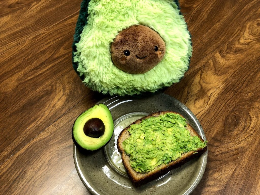 Cholesterol Blog insert image. A plate with a piece of cholesterol friendly avocado toast and half of an avocado. A cute Avocado plush doll (yes a plush avocado with a face) from Squishables sits above the plate.