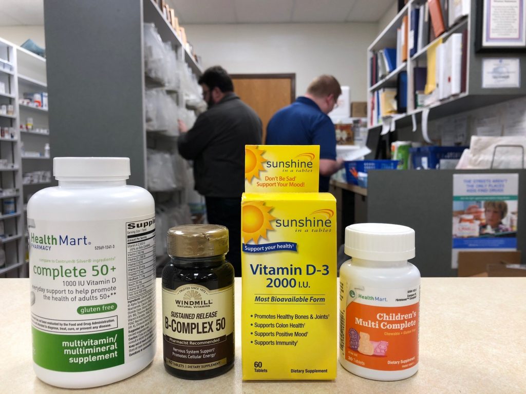 Vitamins on the pharmacy counter at Oswald's Pharmacy. A multivitamin, B-complex vitamin, D vitamin and children's vitamin selection. Two employees can be seen working in the background.