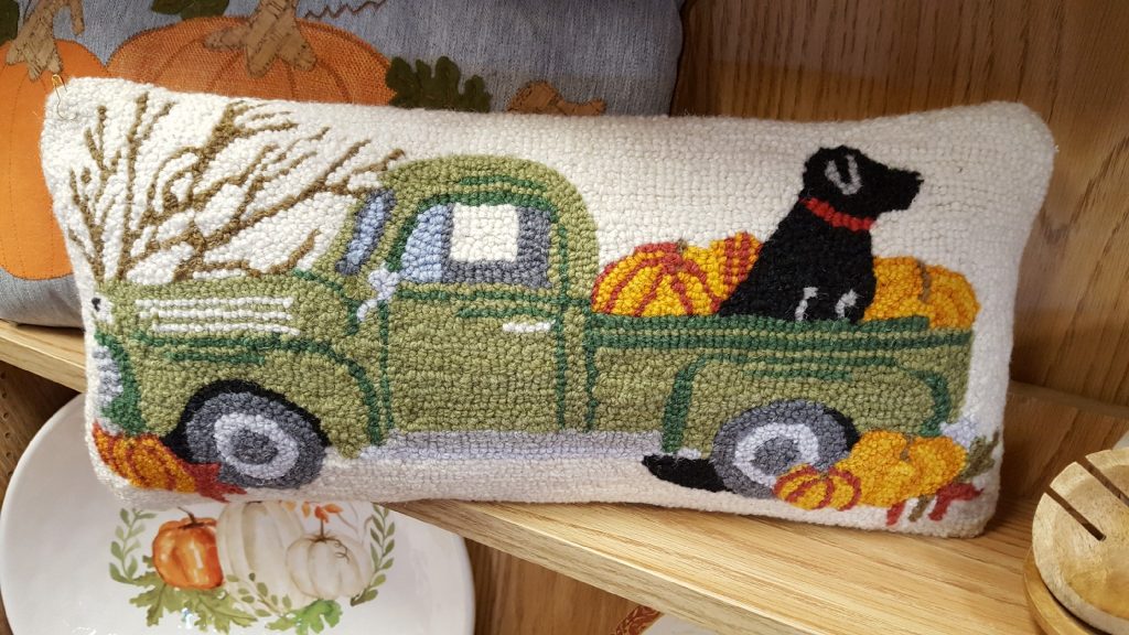 Mud Pie French Knot Pillows Image. Just like grandma used to make! A pillow sitting on the shelves in Oswald's Holiday section picturing a black lab riding in a green pickup truck filled with pumpkins.