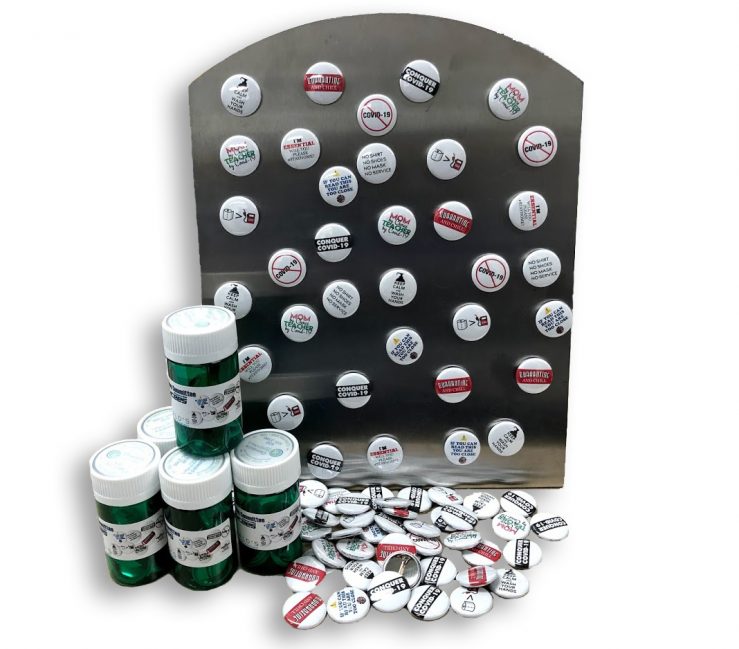 COVID-19 Pins and Magnets display. A picture of a black magnet display with the Oswald's Pharmacy COVID-19 relief pins stuck on it. A pile of buttons with the same images and packaged buttons are in front of the magnet display.