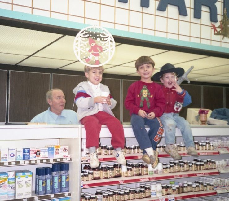 A photo of the pharmacy counter at Oswald's Pharmacy in 1990. 4th generation owner Robert Anderson is standing behind the counter working as the pharmacist for the day. 3 of his grandchildren (Wil, Alex, and Max) are seated on top of the pharmacy counter for the photo with grandpa.