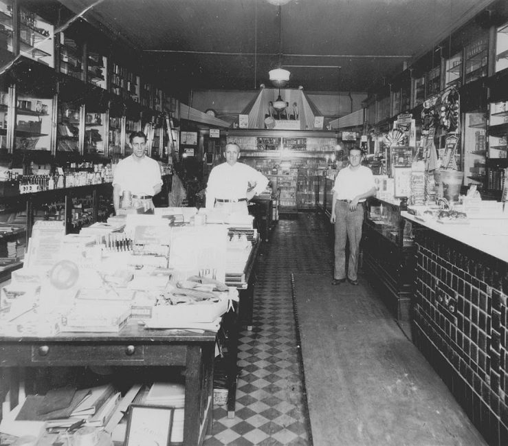 A photo of the interior of Oswald's Pharmacy in 1930. The black and white photo shows a traditional pharmacy. Louis Oswald and Harold Kester (2nd and 3rd generation owners, respectively) are standing in the background with a few other employees.