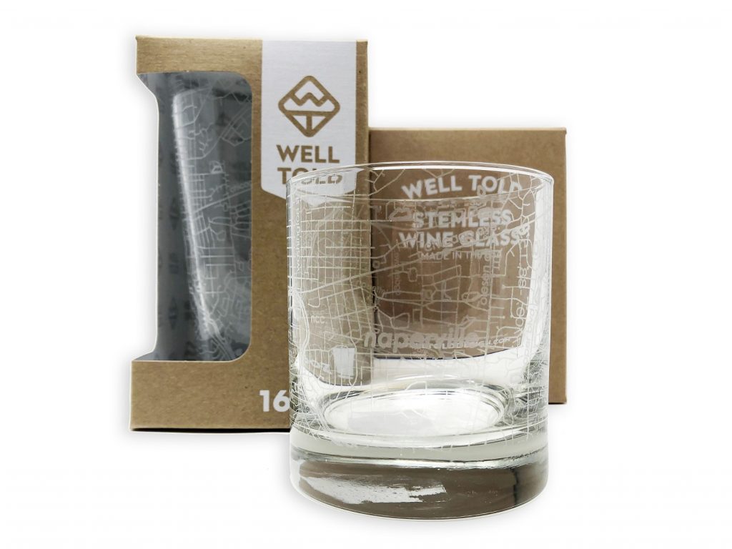 Well Told Naperville Glasses. Photo of a highball glass with Naperville road maps etched into it. Background shows a 12oz glass with similar etchings still in packaging.